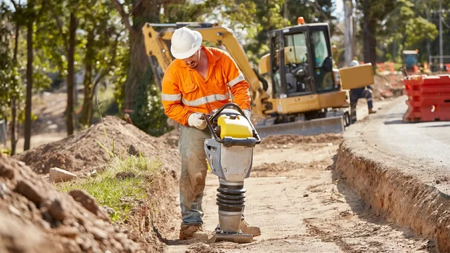 A worker compacting ground beside an excavator